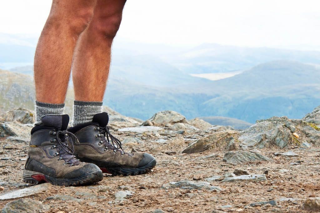 A gay man's thick legs showing off his well-worn hiking boots and a vast countryside in the background.