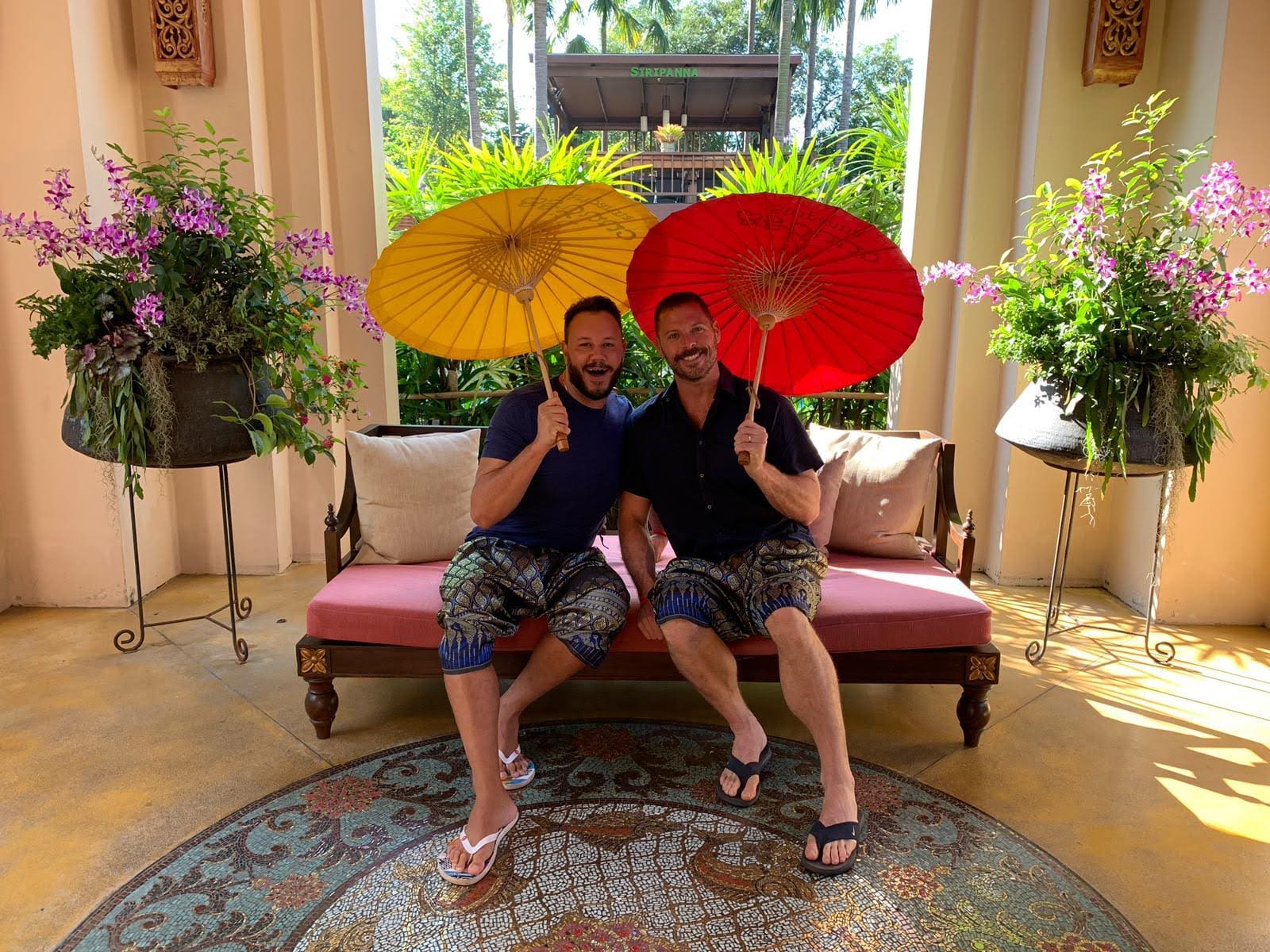 Two gay travellers in Thailand wearing traditional clothes and holding umbrellas.