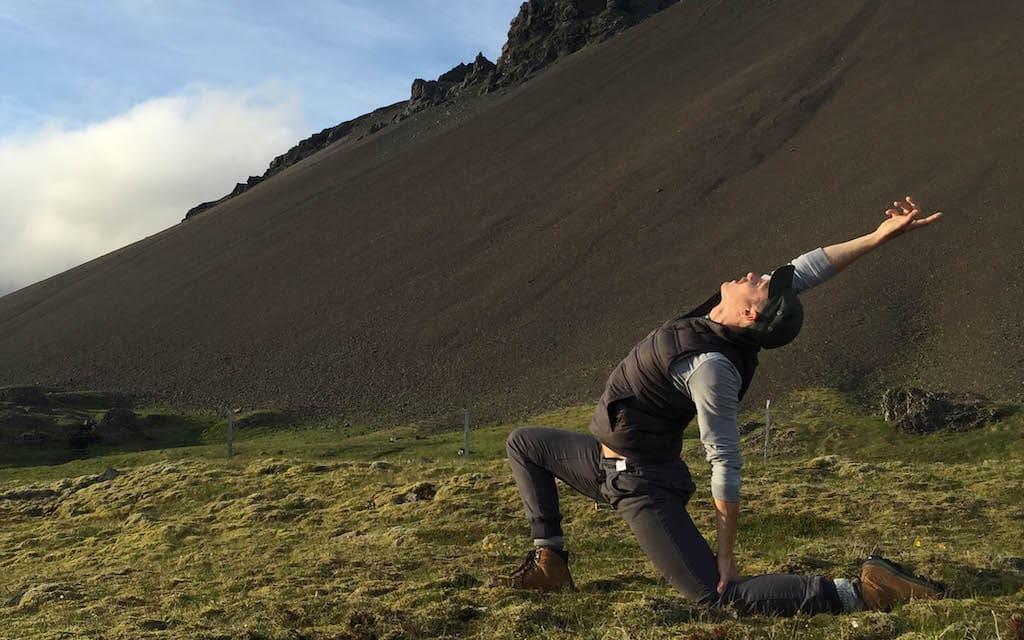 Adam Bolton enjoys a deep lunge during a gay yoga retreat with a hilly landscape in the background.
