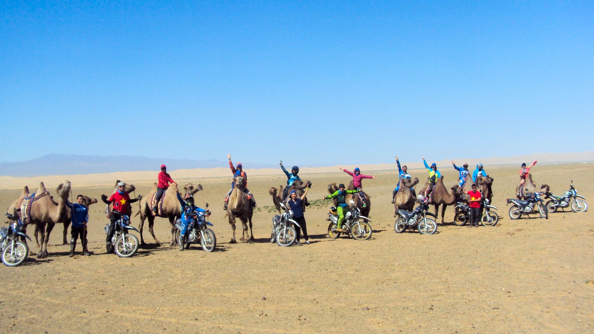 A group of travellers in Mongolia's Gobi Desert wave at the camera. Some are on camels while others are on motorcycles.